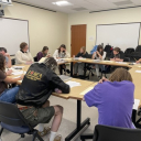 A group of students sit at tables arranged in a large circle and closely examine paper documents.