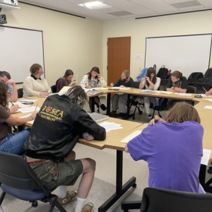 A group of students sit at tables arranged in a large circle and closely examine paper documents.