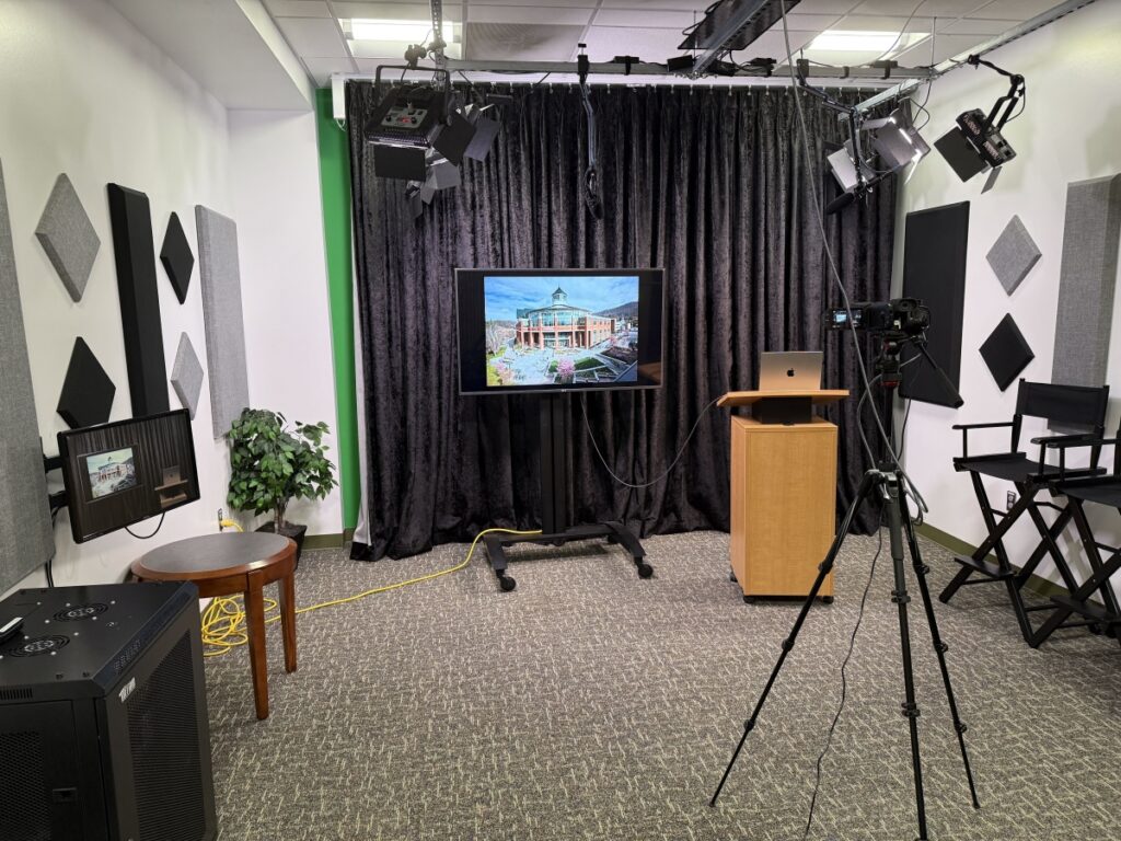 Recording studio with black curtain backdrop and a display screen connected to a laptop on a podium